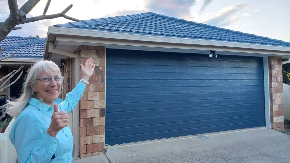 A smiling woman wearing a blue shirt and glasses gives a thumbs-up while standing in front of a house with a newly installed blue garage door and a clean roof